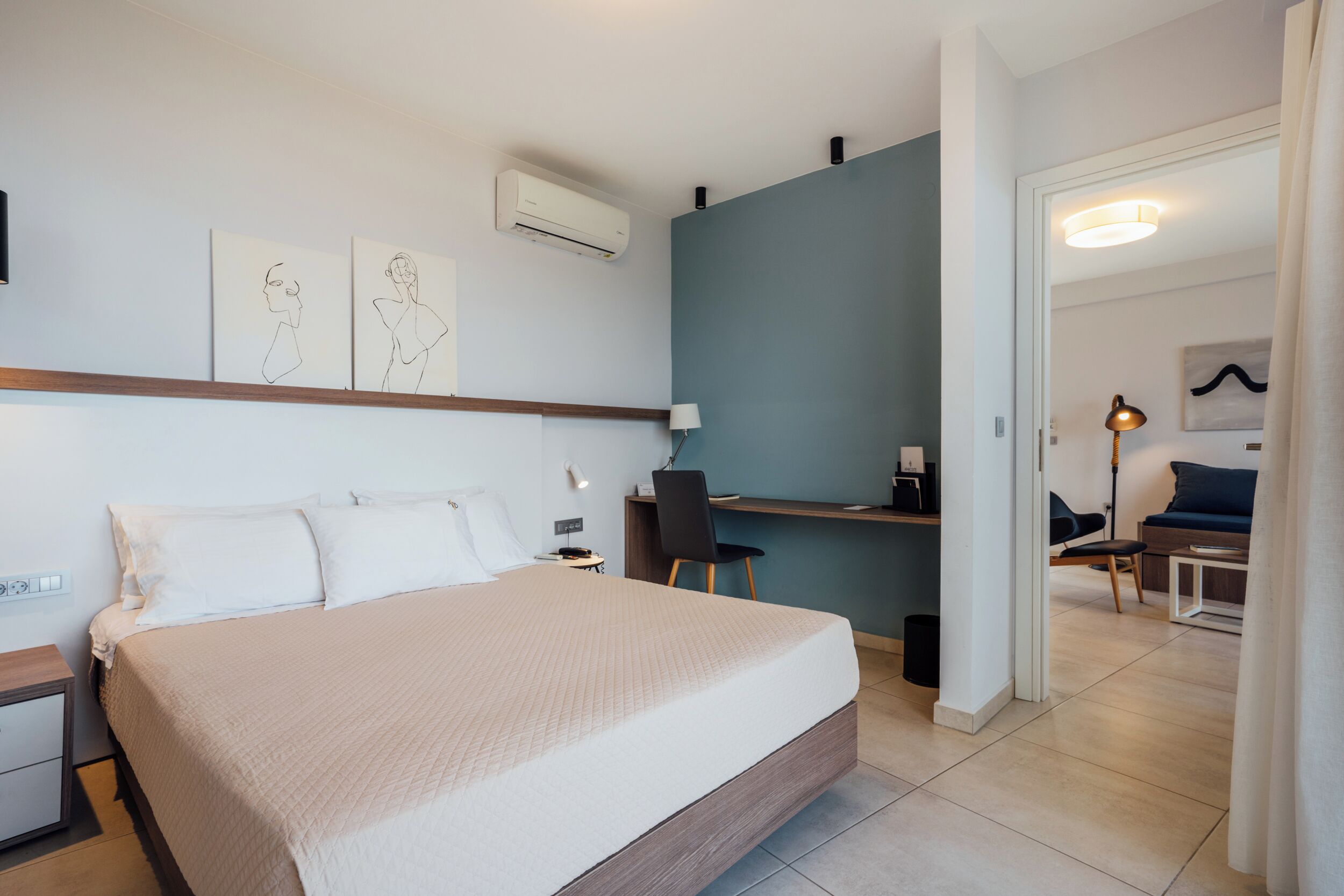 A modern hotel room with a large bed, a desk, and a chair. There is a wall-mounted air conditioner and a lamp on the desk.
