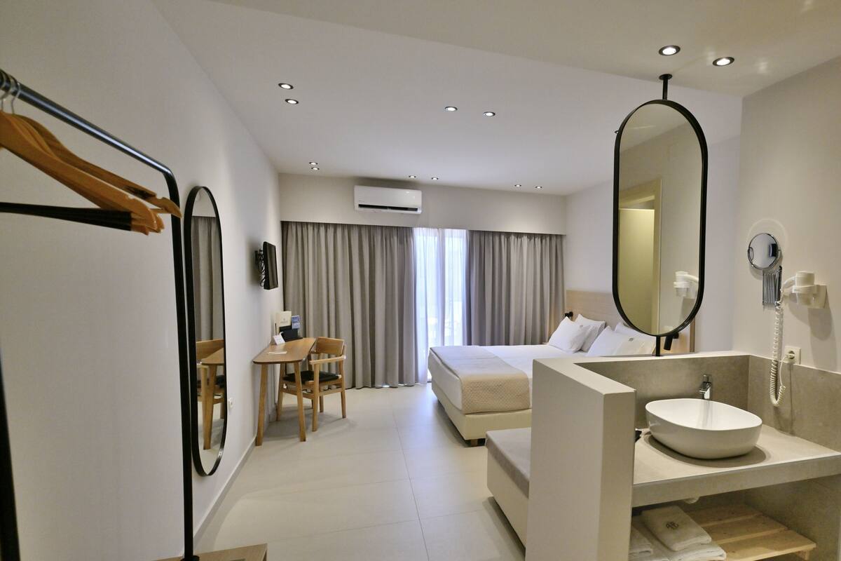 A modern hotel room with a bed, a desk, a chair, a bathroom with a sink and mirror, and an air conditioning unit.