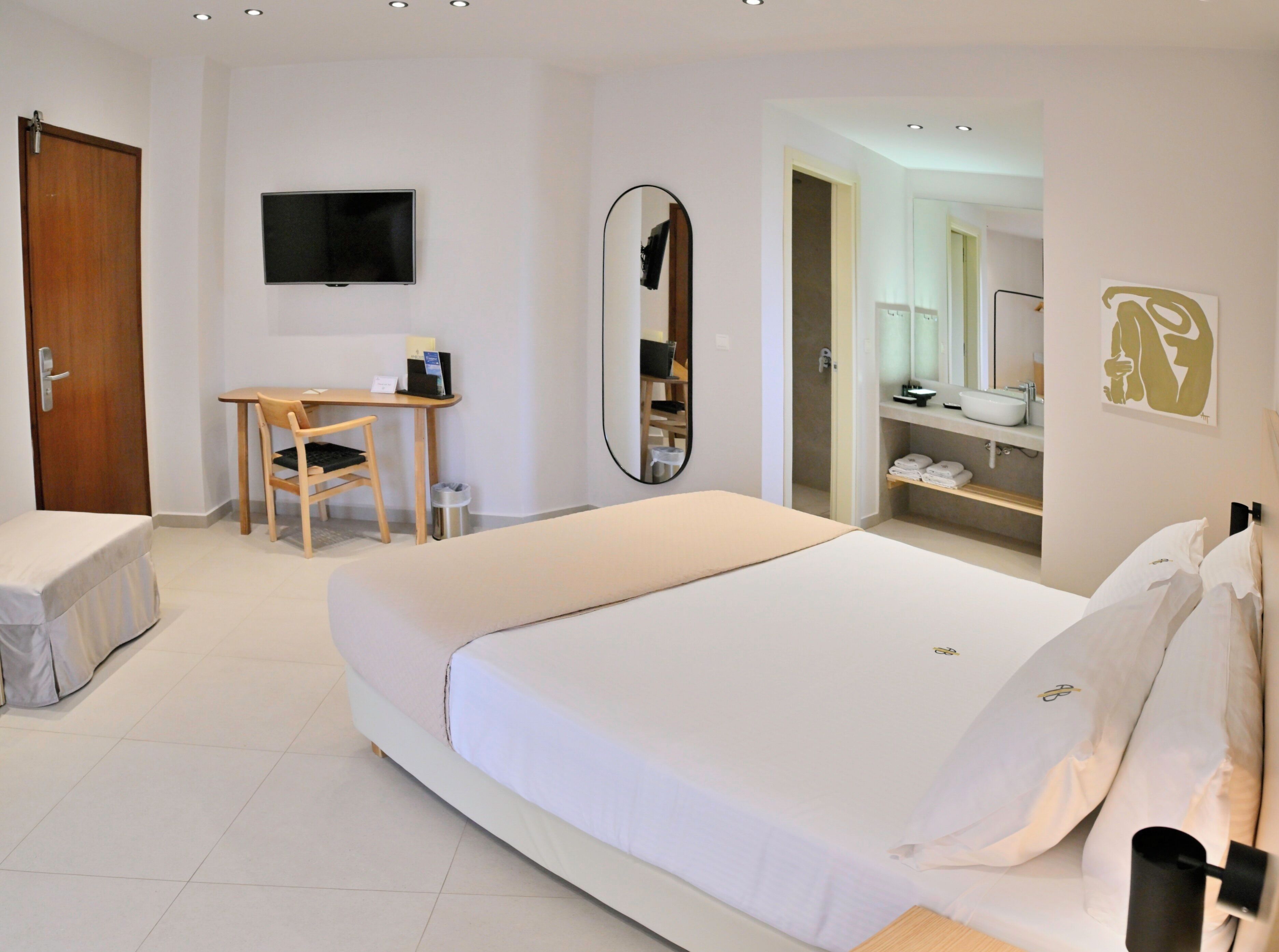 A hotel room with a bed, a desk, a chair, a TV, a mirror, and a bathroom visible through an open door.