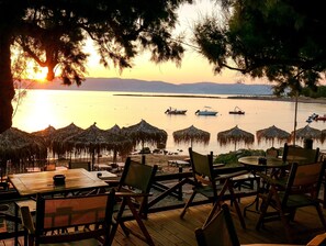 A beachside restaurant with thatched umbrellas and wooden deck seating overlooking calm waters and distant mountains. - Aphrodite Beach (Kissamos)