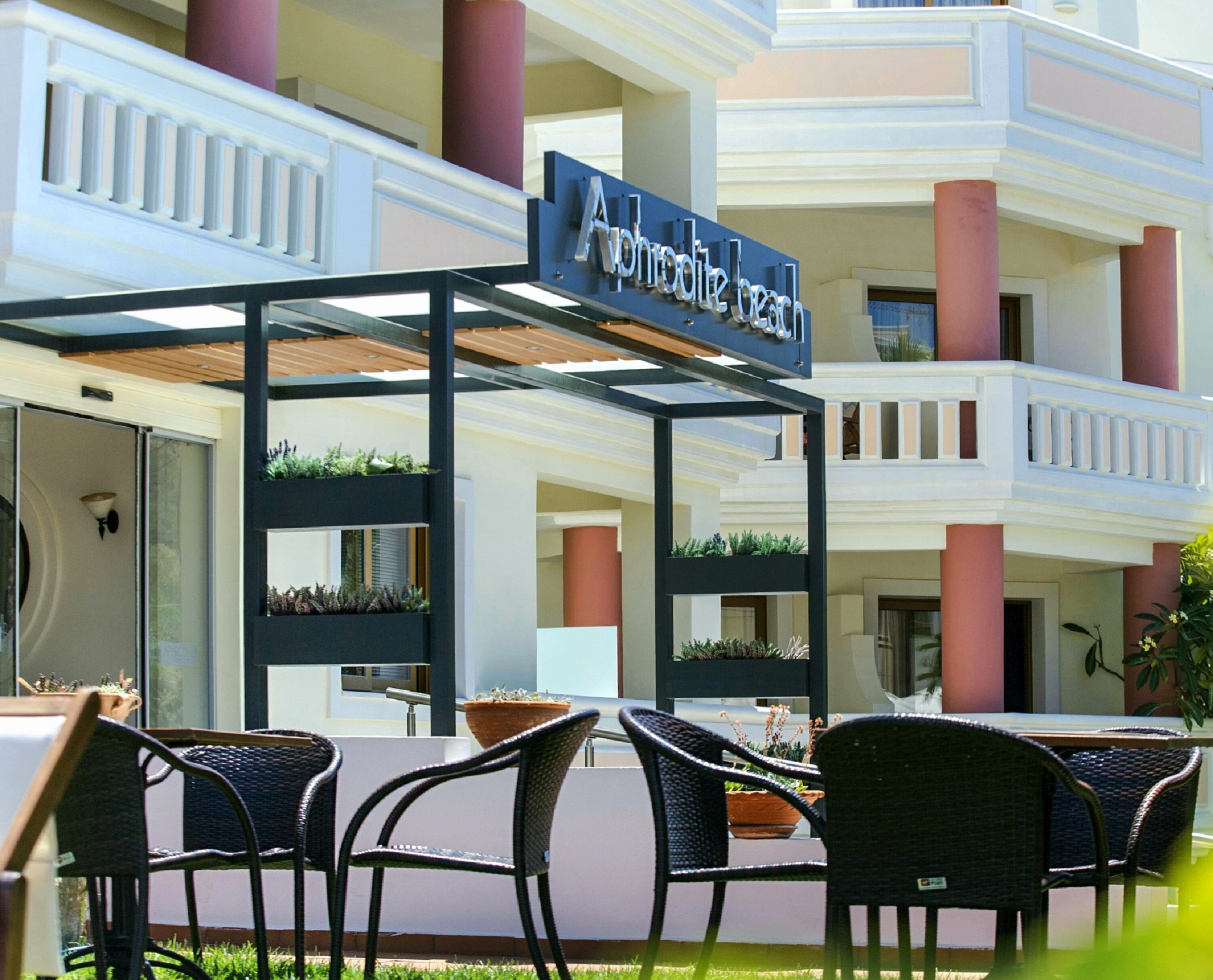 Un vestíbulo de hotel con asientos exteriores, un letrero que dice "Aloha Beach" y varios balcones con columnas rojas.