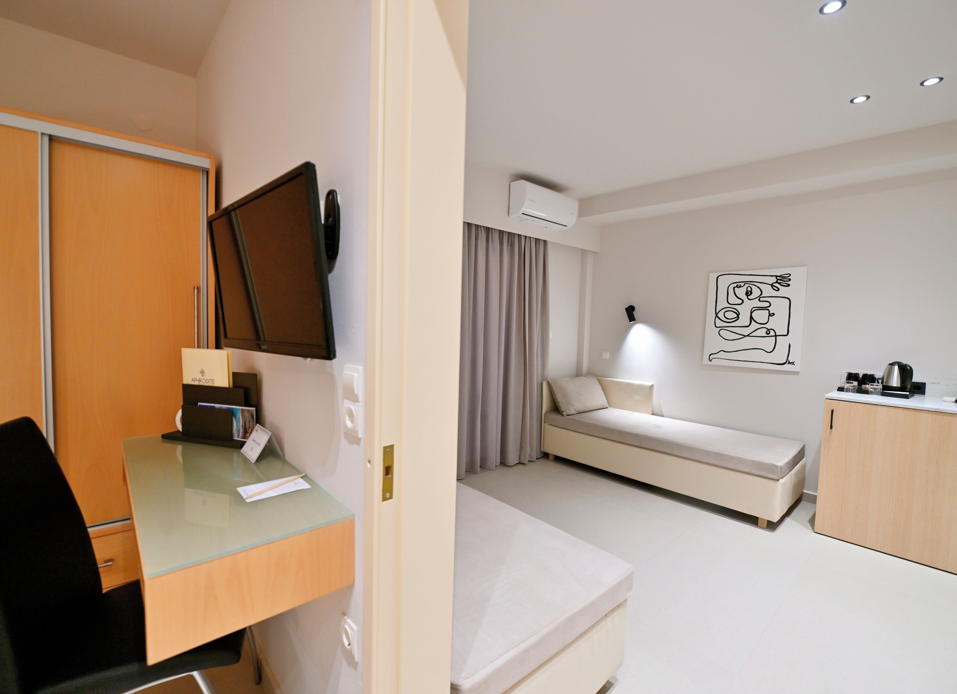 A modern hotel room with a desk, a bed, a small sofa, a coffee machine, and a painting on the wall.