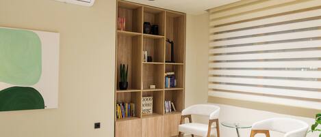 A modern living room with a wooden bookshelf, two white chairs, and a large window with blinds.