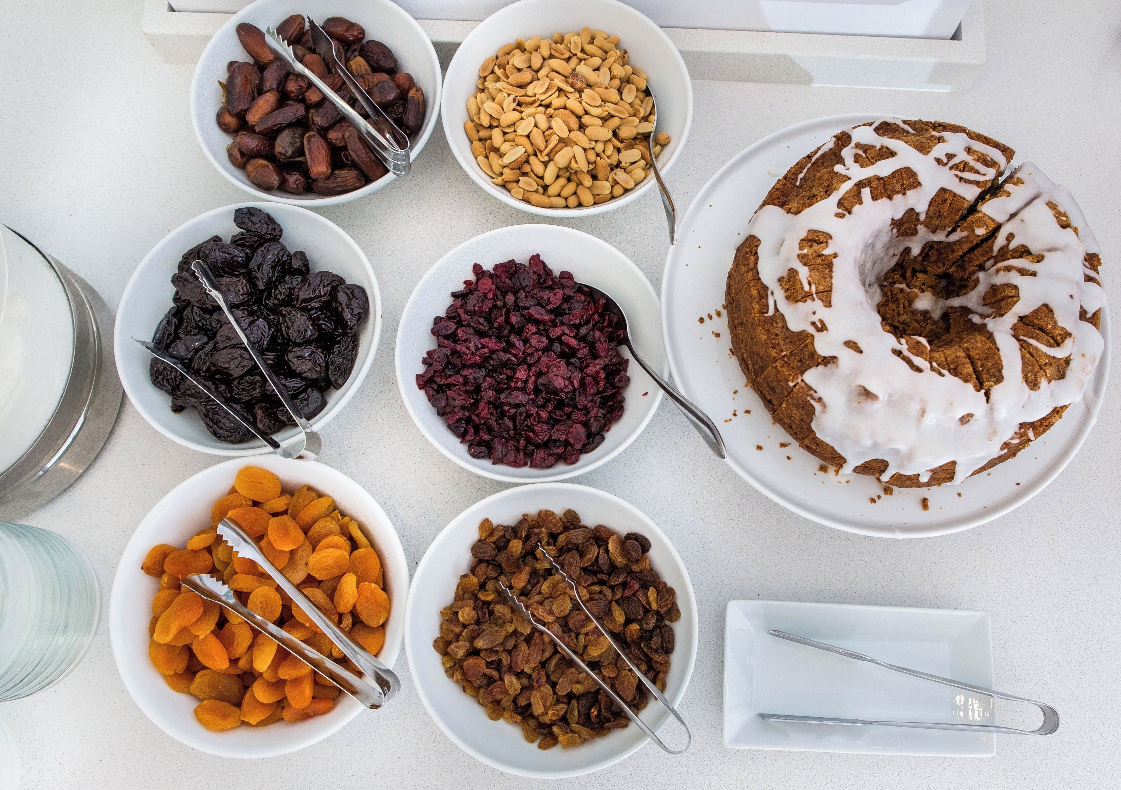 A table with various bowls of nuts and dried fruits, a cake with white icing, and a glass of milk.