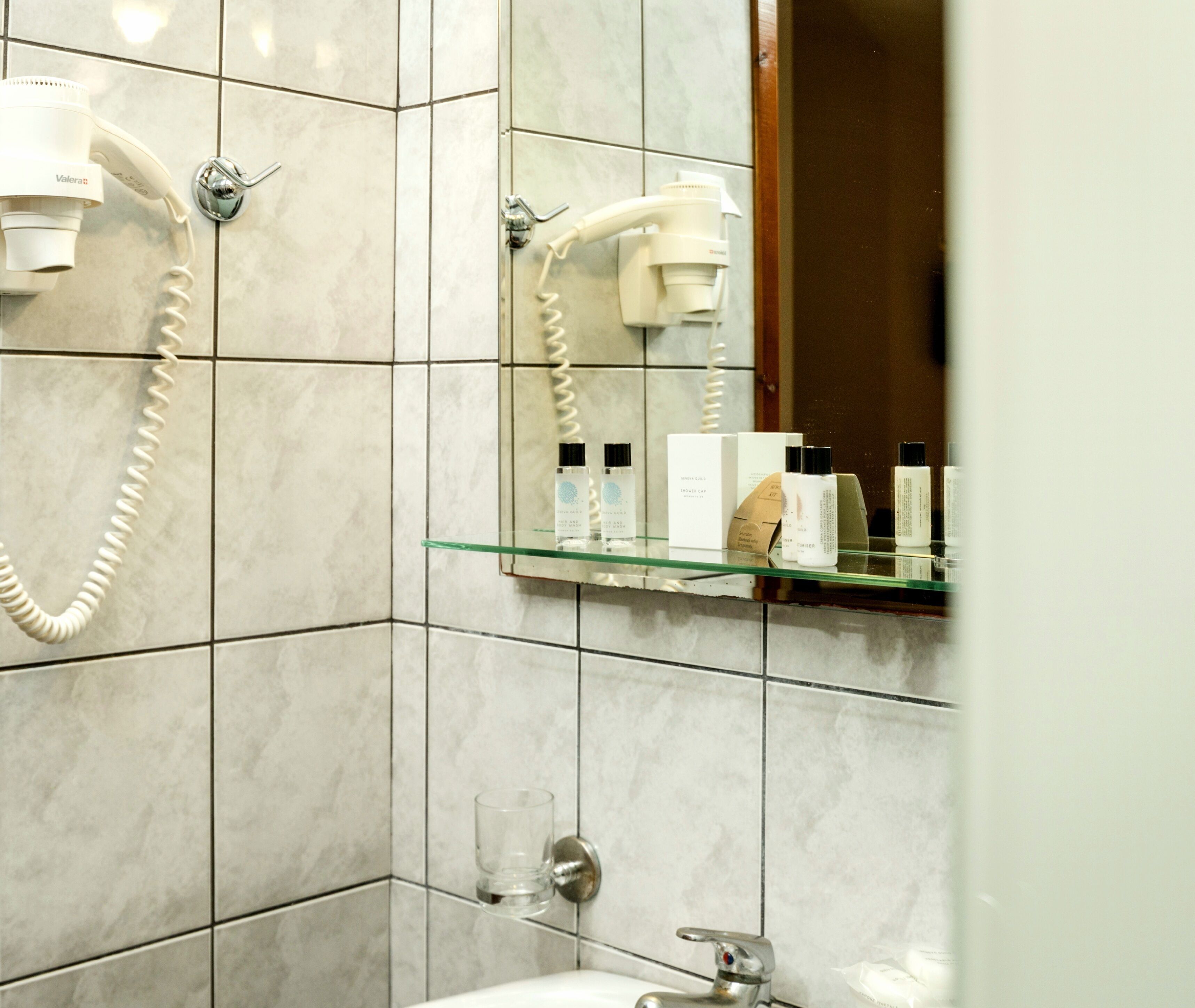 Une salle de bain équipée d’un sèche-cheveux fixé au mur, d’une étagère en verre où sont rangés les articles de toilette, et d’un miroir qui reflète le sèche-cheveux.