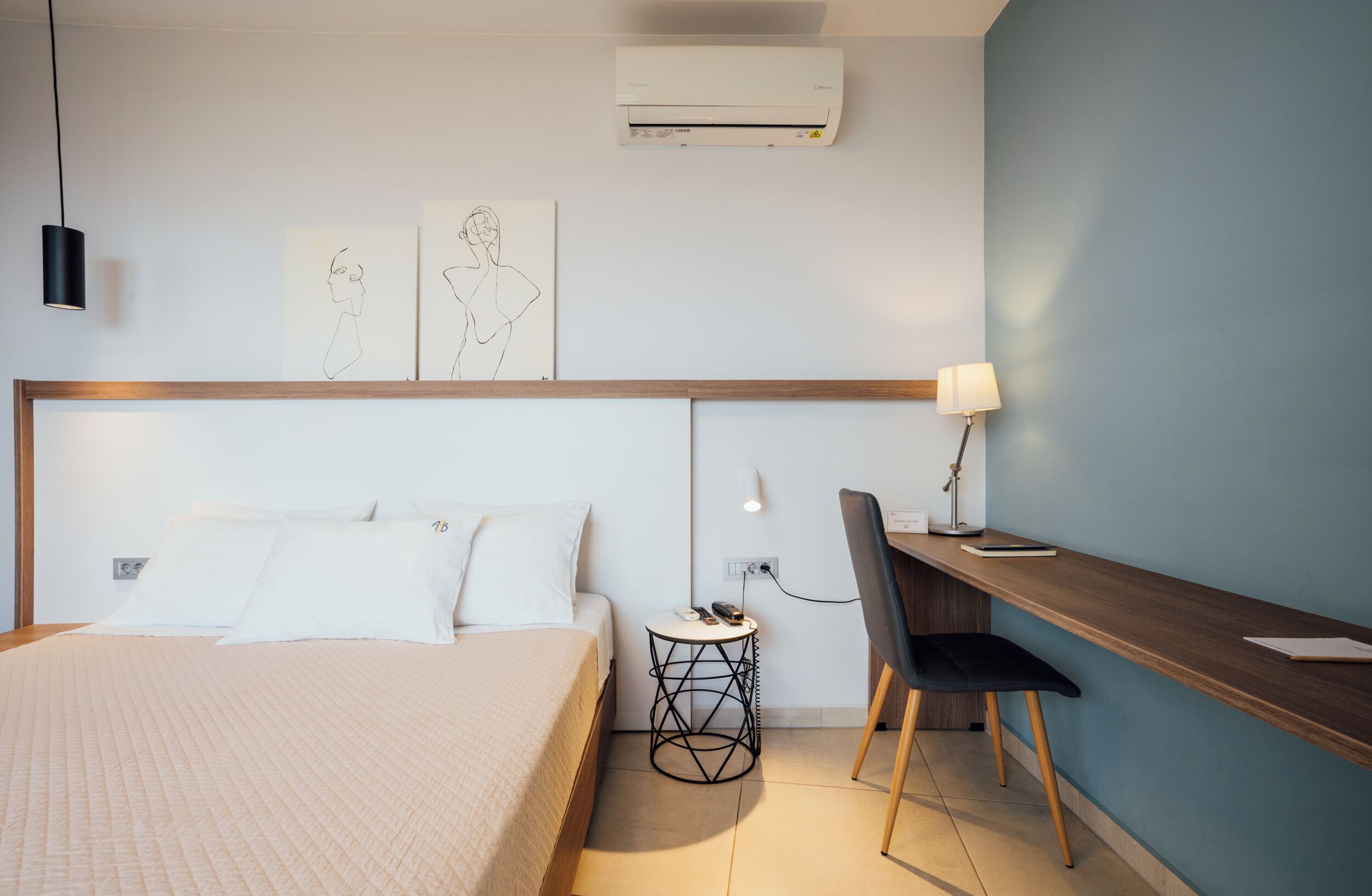 A modern hotel room with a bed, a desk, and a chair. There is a wall-mounted air conditioner and a small side table with a lamp.