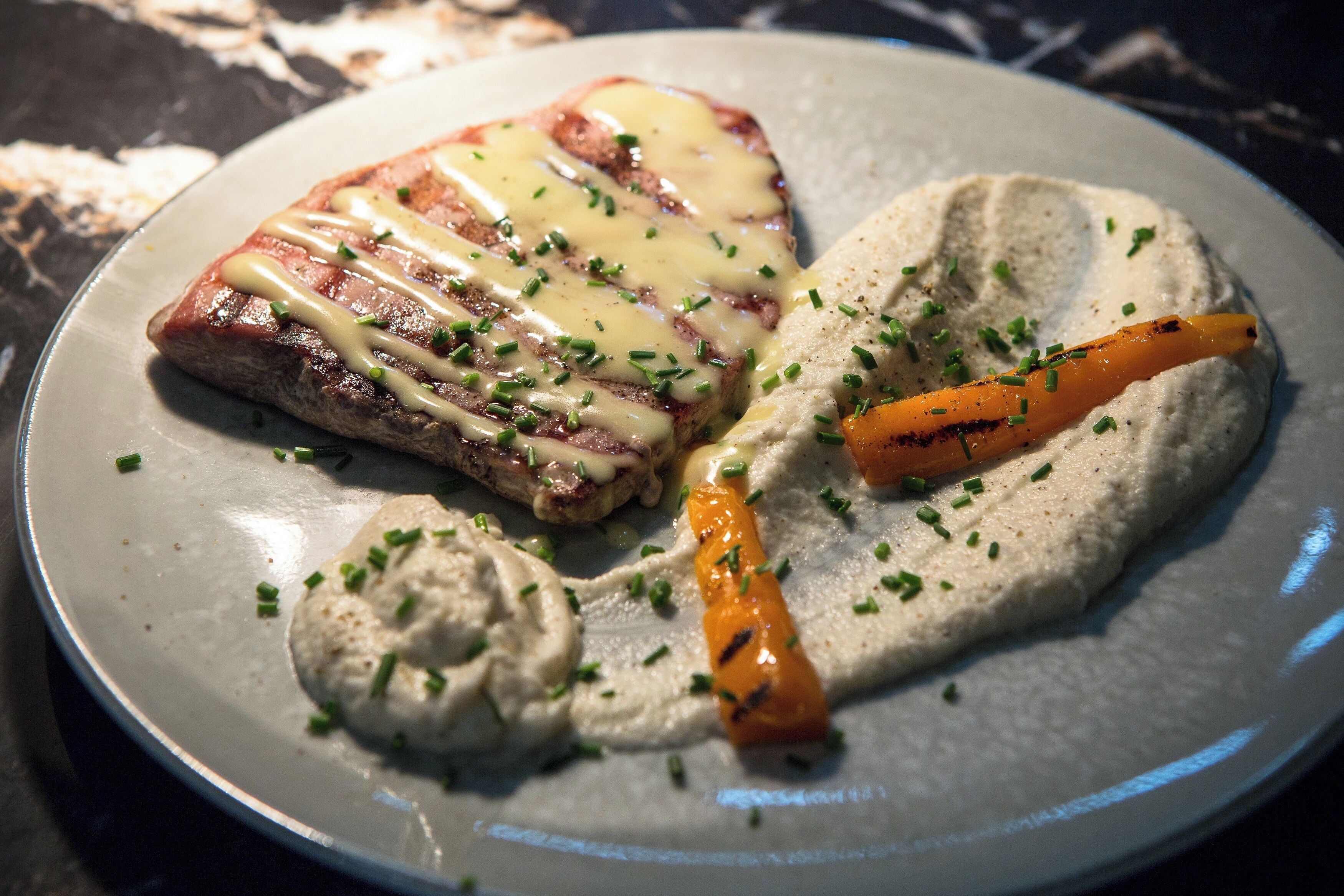 A plate with a grilled steak, a slice of cheese, a grilled carrot, and a dollop of hummus.