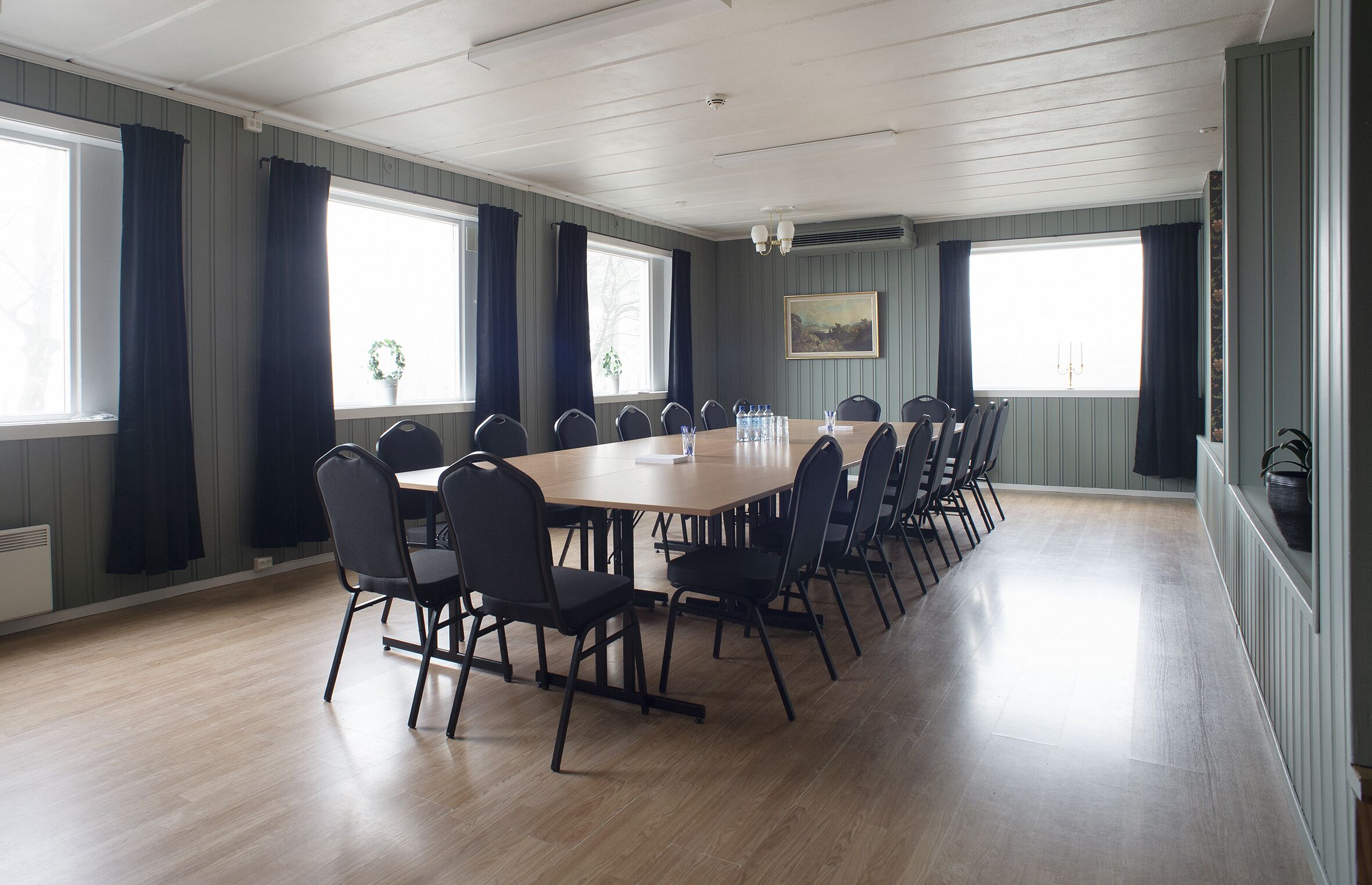 A conference room with a long table and chairs, large windows with curtains, and a painting on the wall.
