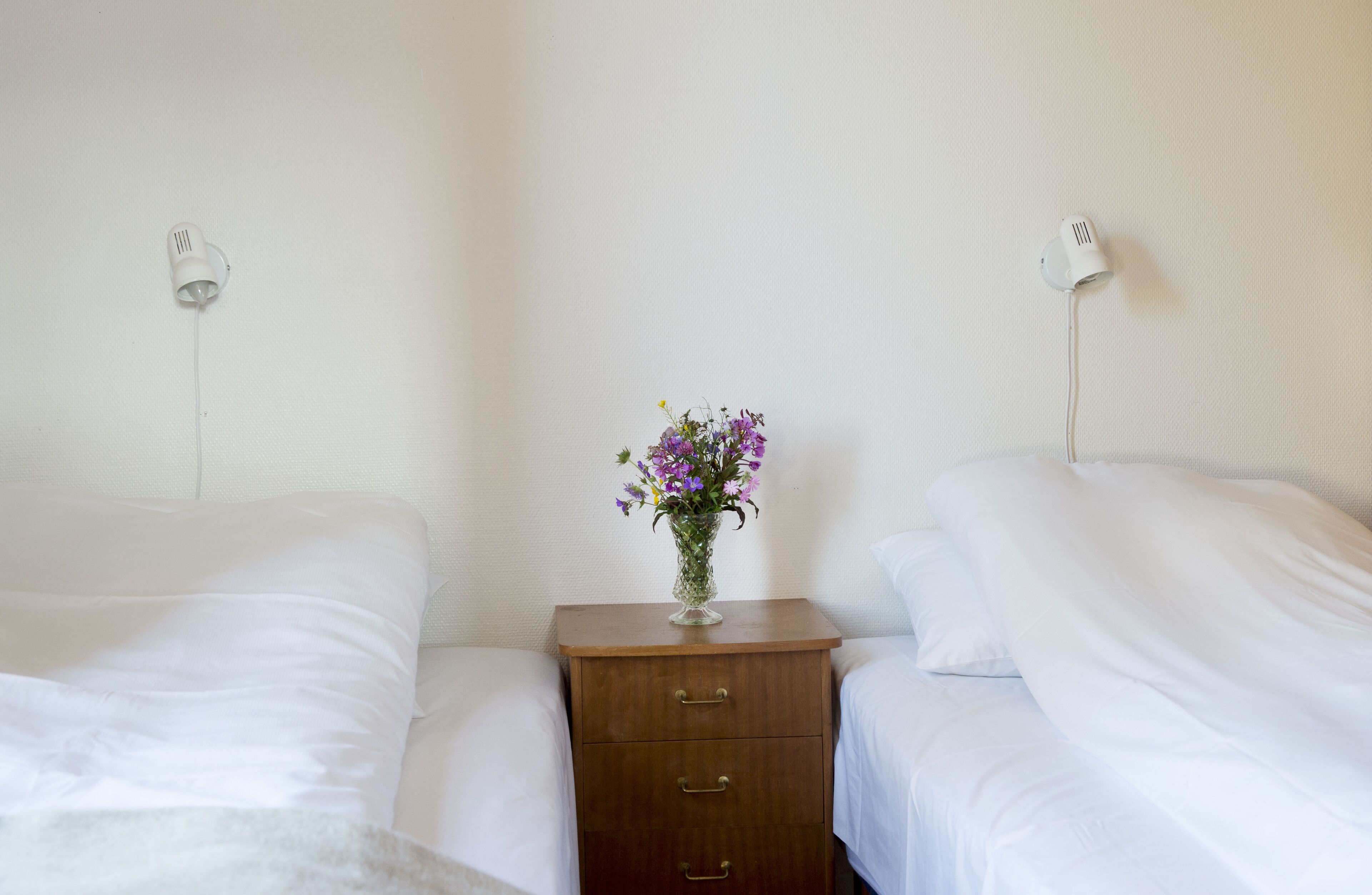 A hotel room with two single beds, a wooden nightstand with a vase of flowers, and wall-mounted lamps.