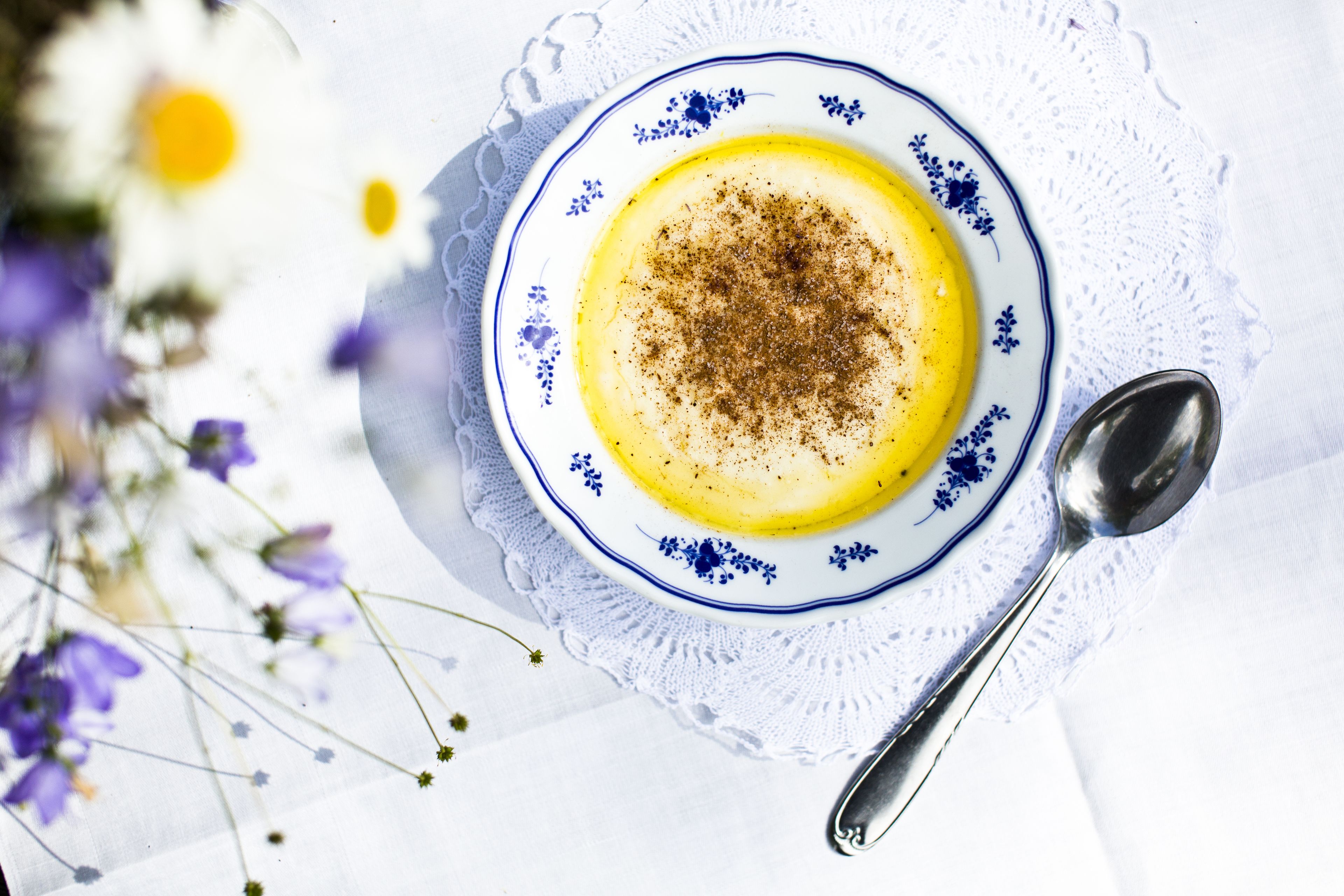 Een kom met geel dessert met sprinkles, geplaatst op een wit bord met blauwe bloemmotieven, samen met een lepel op een kantklosje.