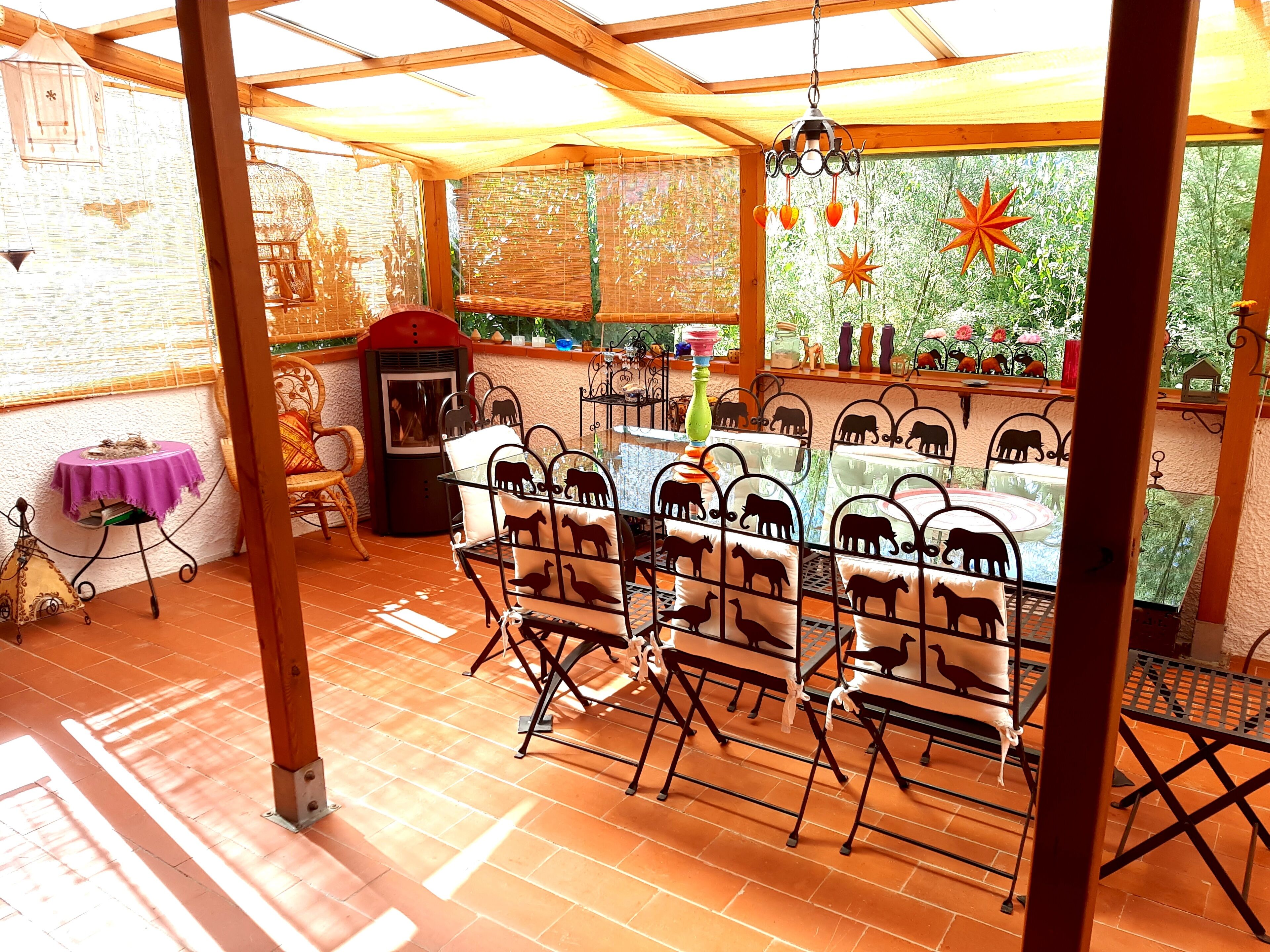A covered outdoor dining area with metal chairs and tables, a brick floor, and a view of greenery through the windows.