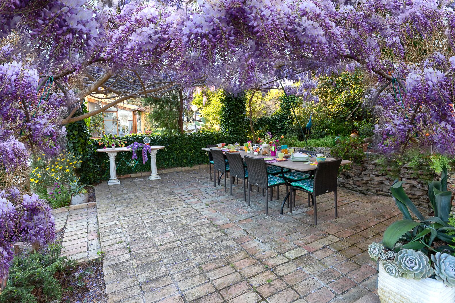 Eine Terrassengruppe im Freien mit einer Pergola, die mit violetten Blüten bewachsen ist, Steinboden und einem Esstisch.