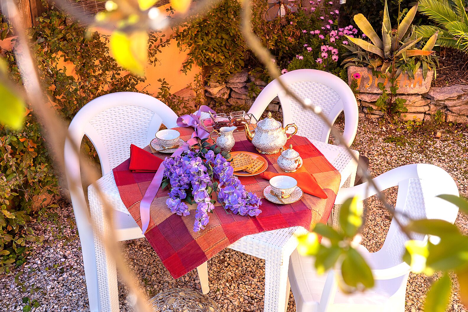 An outdoor dining area with a table set for two, featuring a checkered tablecloth, white chairs, and a floral centerpiece.