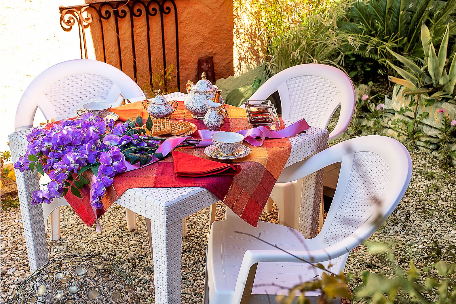A table set outdoors with white wicker chairs, a red and orange tablecloth, and purple flowers.