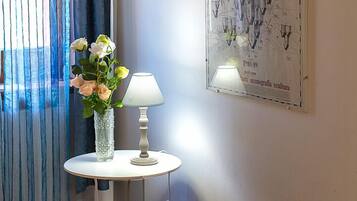 Una habitación con una cama de madera, una mesa blanca con una lámpara, un jarrón con flores y un cartel en la pared.