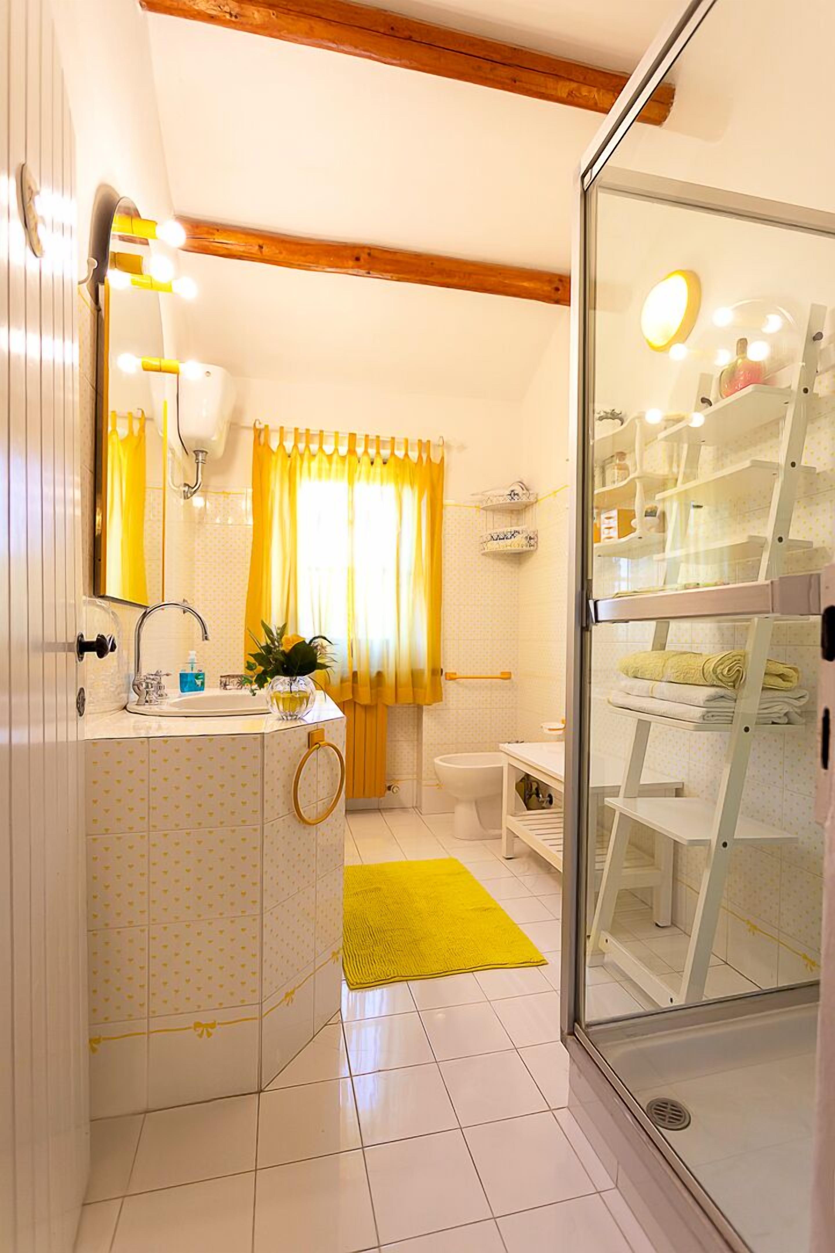 A bathroom with a glass shower enclosure, a white sink with a mirror, yellow curtains, and a yellow rug.