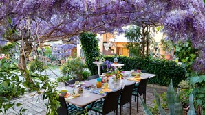 Un área de comedor al aire libre con una mesa dispuesta para cuatro personas, rodeada de follaje exuberante y glicinas moradas.