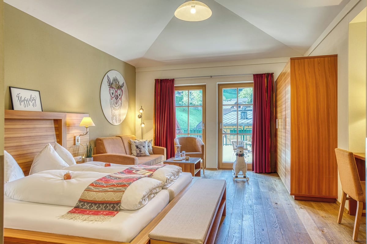 A hotel room with a bed, a sofa, a desk, and a wardrobe. There is a window with curtains and a view of the outside.