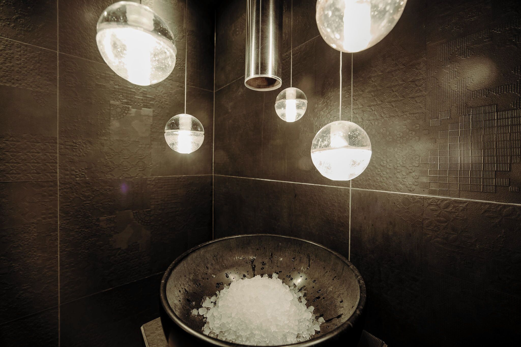 Un baño con un bol de hielo, paredes de azulejos oscuros y luces esféricas colgantes.