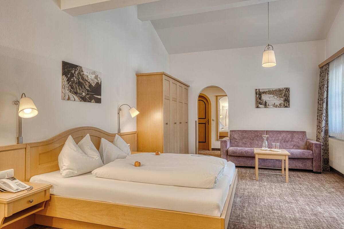 A hotel room with a bed, a wooden wardrobe, a sofa, a small table, and two wall-mounted lamps.