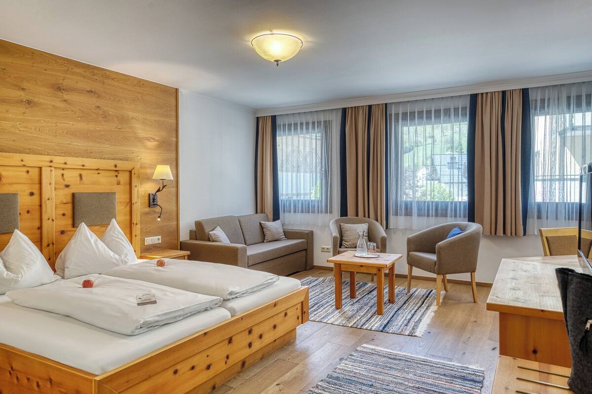 A hotel room with a wooden headboard, a bed with white linens, a sofa, a chair, a small table, and a desk.