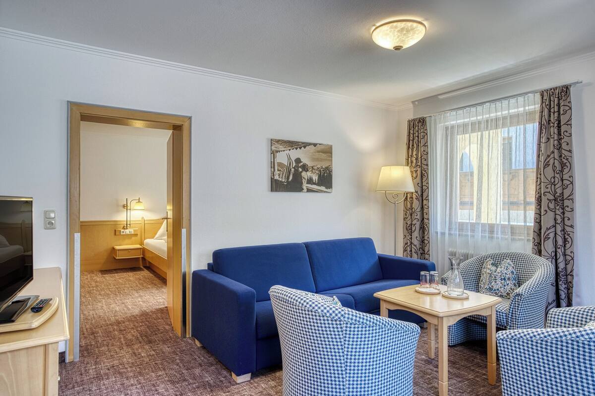 A hotel room with a blue sofa, checkered chairs, a small table with a lamp, a painting on the wall, and a view through sheer curtains.