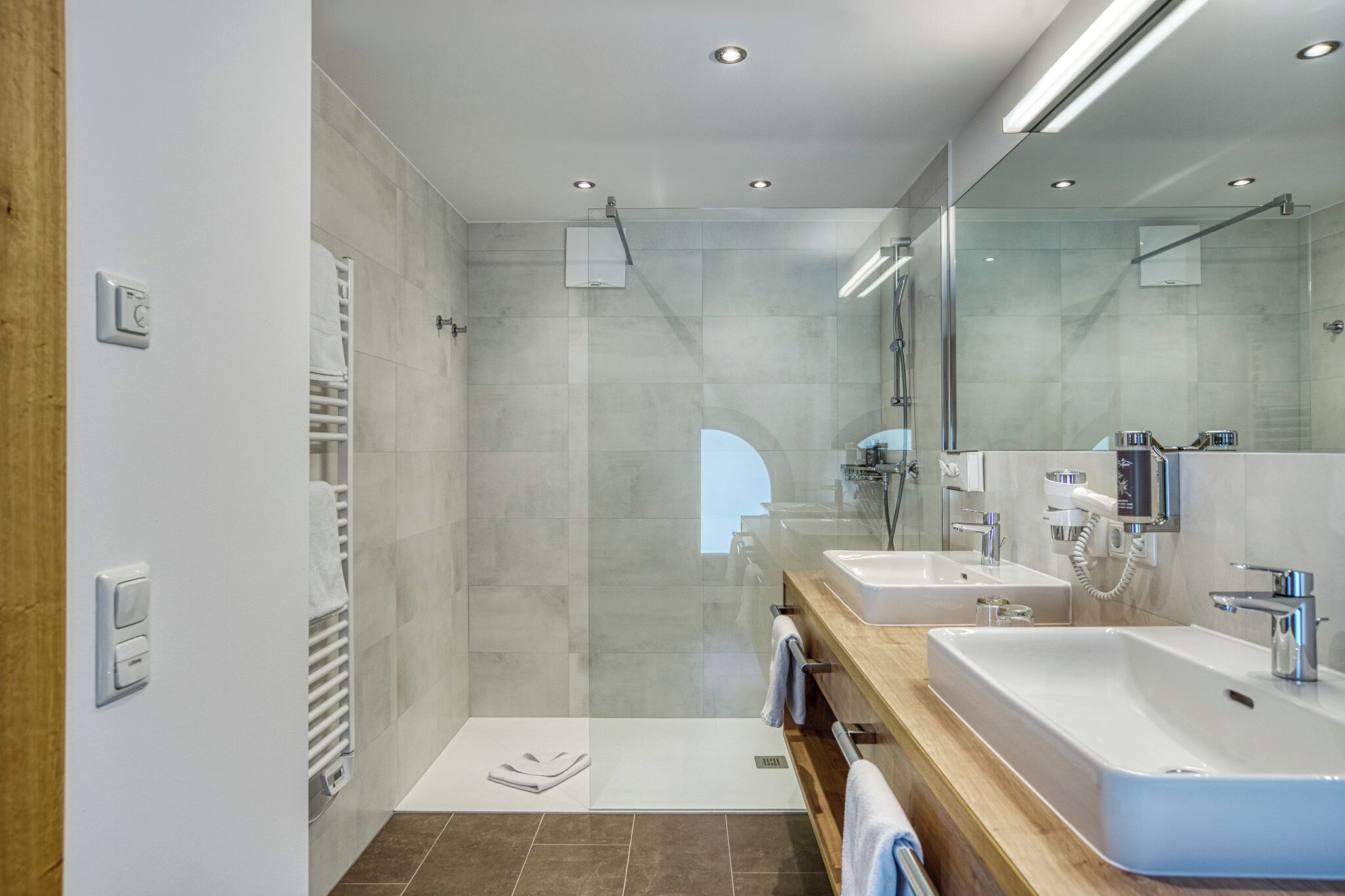 Un baño moderno con cabina de ducha de vidrio, dos lavamanos, un espejo grande y un perchero para toallas.