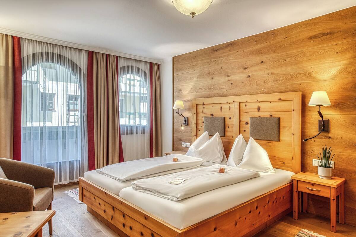 A double bed with white linens, a wooden headboard, two bedside lamps, a small bedside table with a plant, and a view of the outdoors through large windows with sheer curtains.