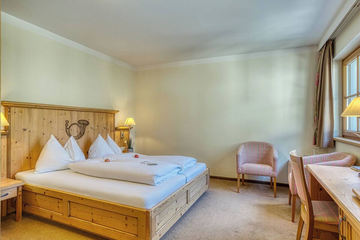 A cozy bedroom with a wooden bed, a small table, a chair, and a window with curtains.