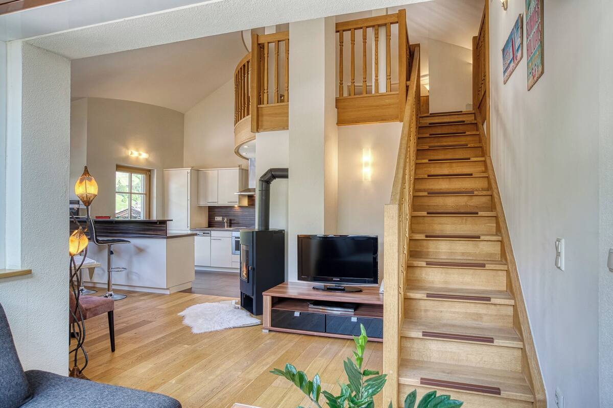 A spacious living area with a wooden staircase, a kitchen in the background, and a flat-screen TV on a wooden stand.