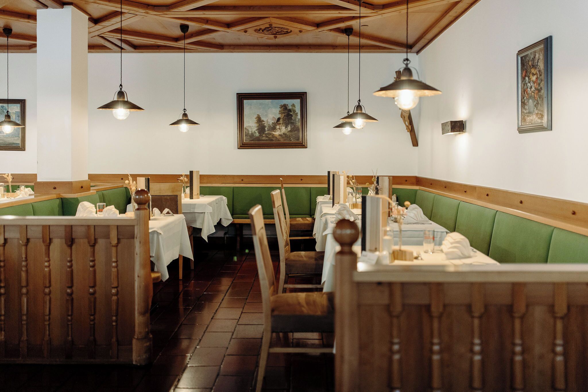 Een restaurant met houten banken, groene gestoffeerde zittingen, witte tafellakens en hangende hanglampen.