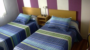 A room with two single beds, each with a striped bedspread and pillows. There is a nightstand with a lamp and a remote control on it. The walls have a striped pattern. - Pensión Gema Irun (Irun)