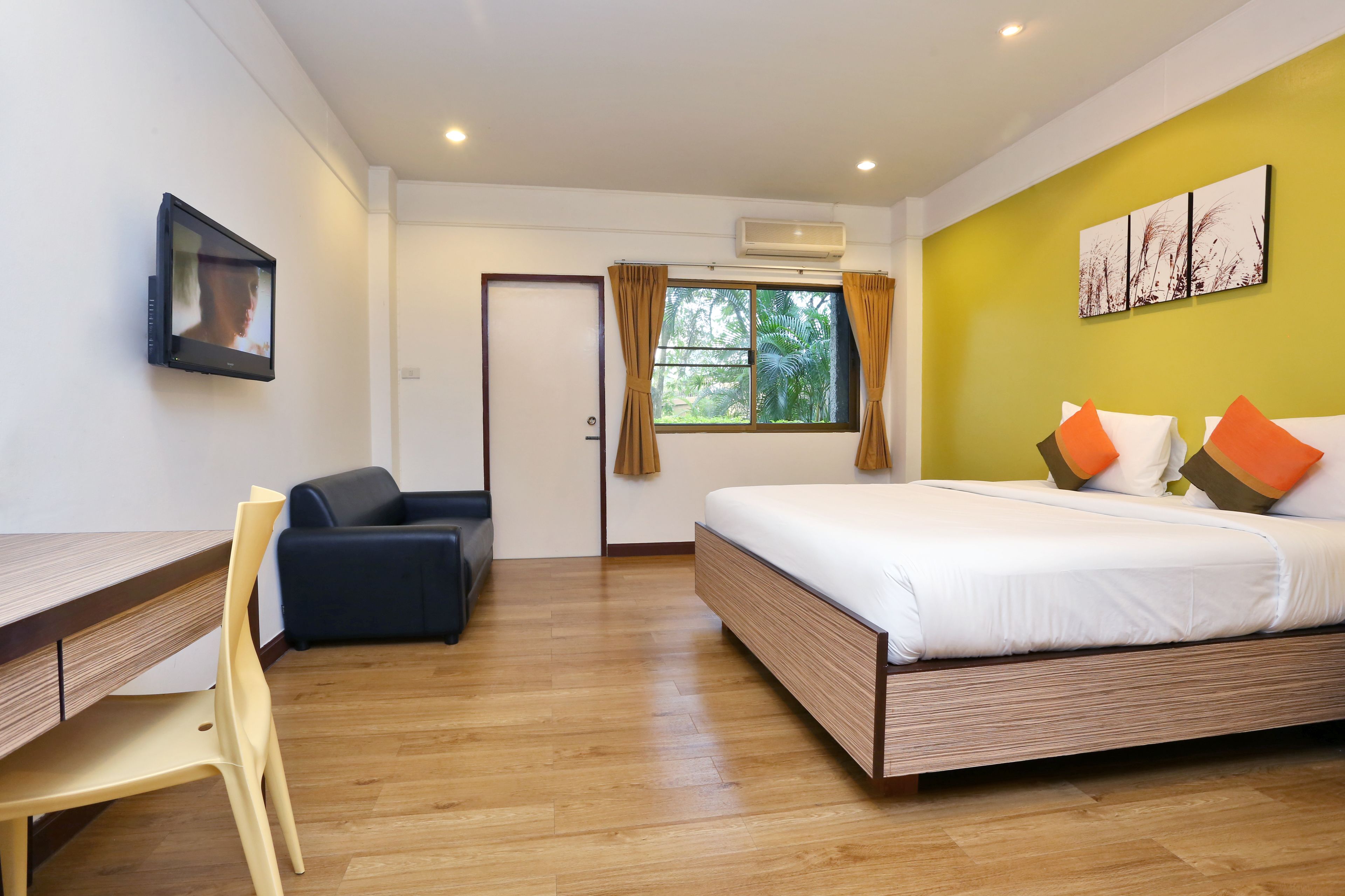 A modern hotel room with a wooden floor, a bed with white bedding and orange pillows, a desk with a chair, a TV mounted on the wall, and a window with curtains.