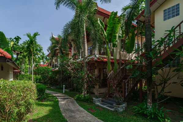 A pathway leading to a building with a red roof, surrounded by lush greenery and palm trees. - Palm Village (Rawai)