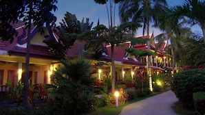 Un hotel de arquitectura tradicional, con varios edificios de techos rojos y un camino que atraviesa un jardín ajardinado con palmeras y otras plantas tropicales.