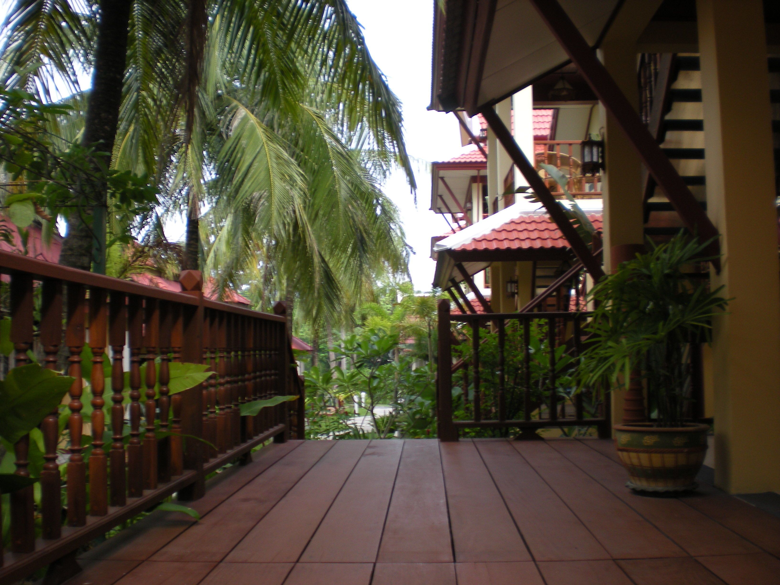 A wooden deck with a railing, surrounded by lush greenery and a red-tiled building with a staircase.