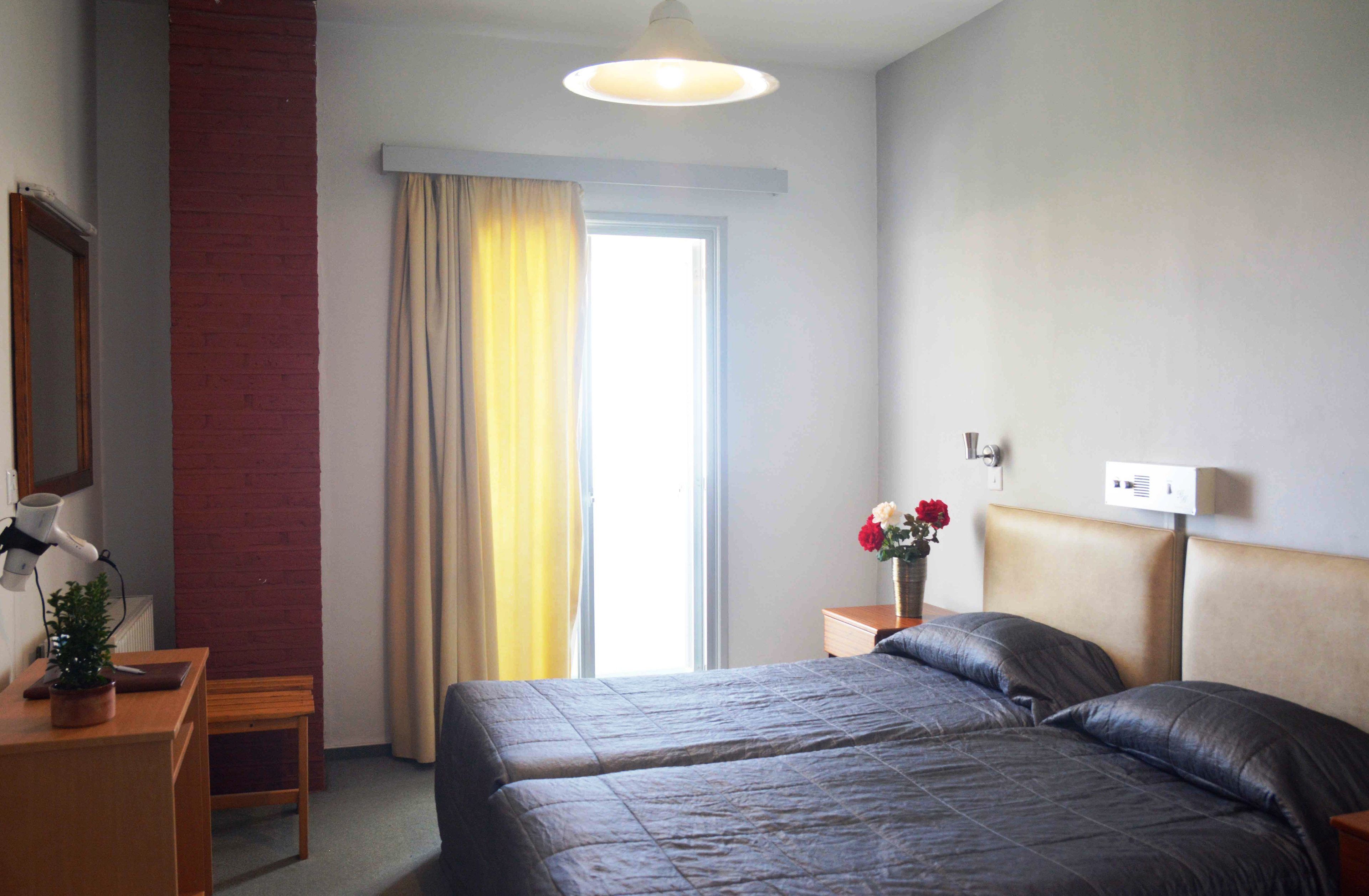 Habitación de hotel con dos camas, una ventana con cortinas, una mesita de noche con lámpara y una planta pequeña.