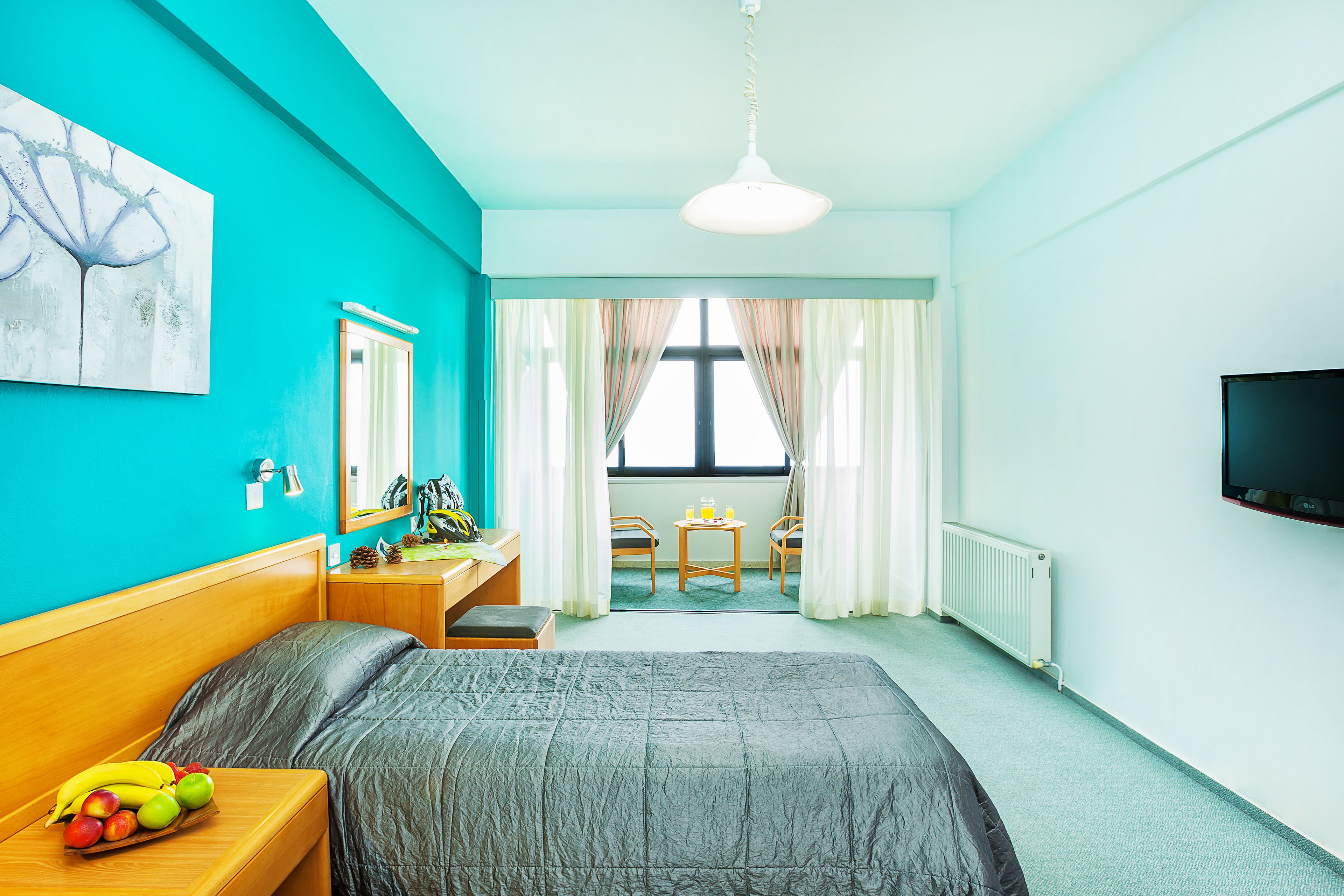 A hotel room with a bed, a fruit bowl, a desk with a mirror, a TV, and a view of the outside through a window.