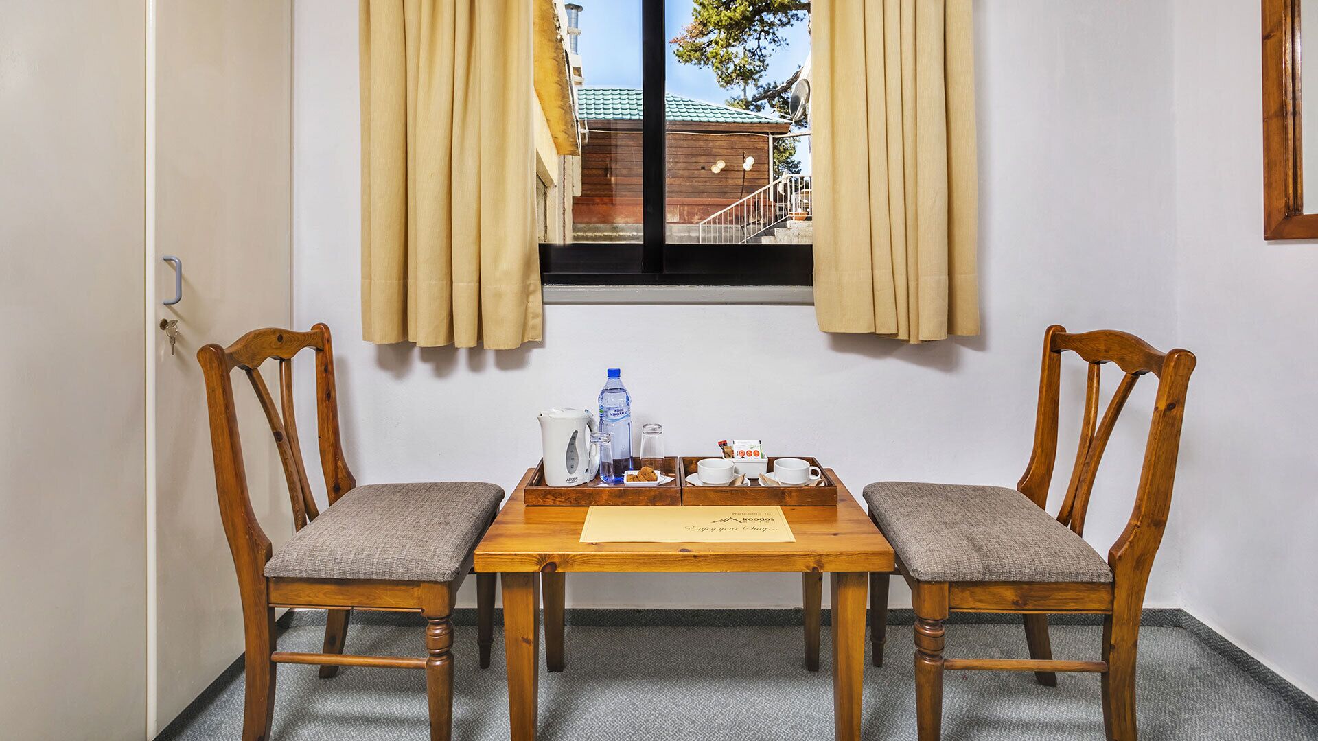 A room with two wooden chairs, a wooden table, a window with curtains, and a view of a building outside.