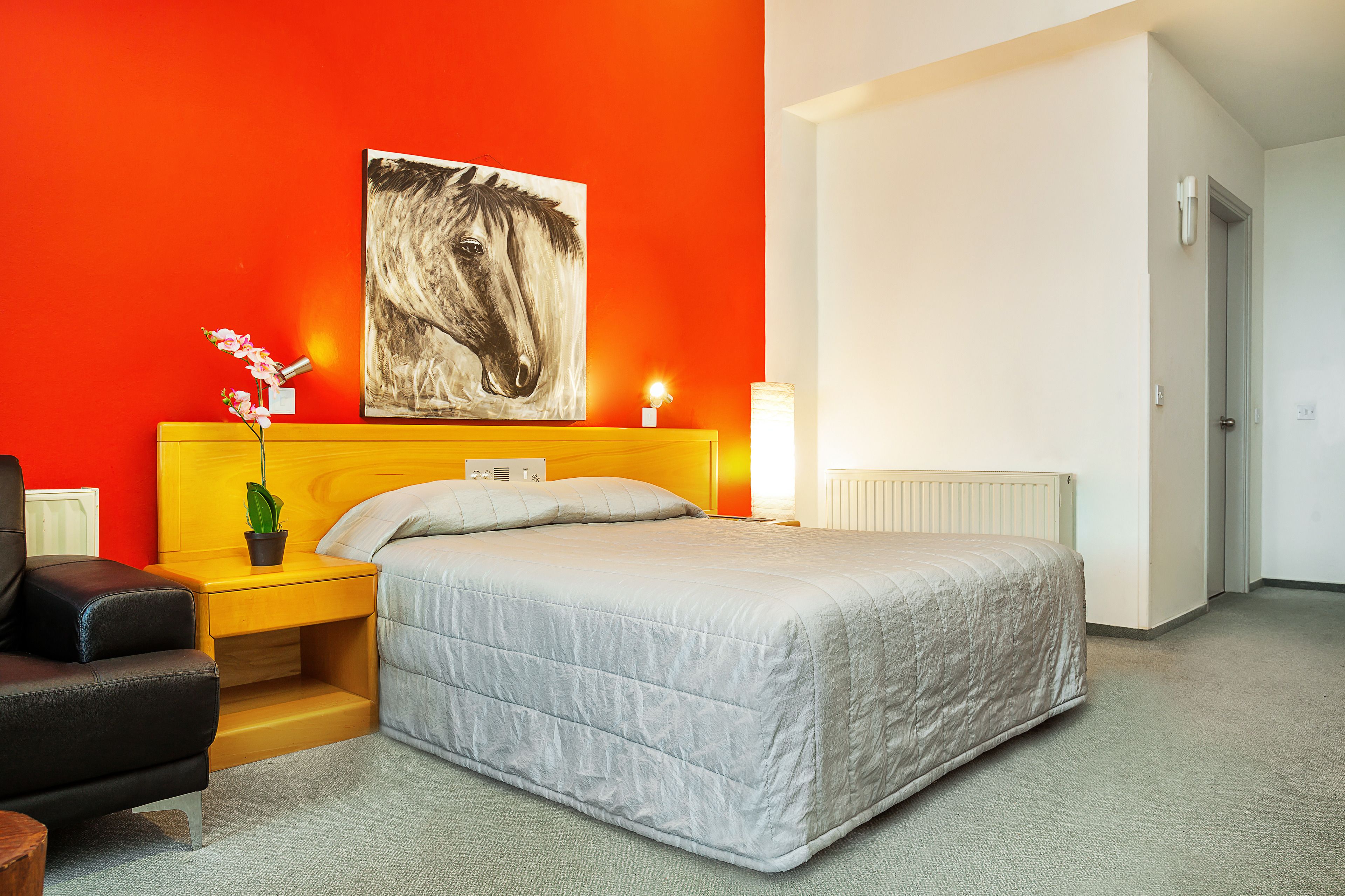 A hotel room with a bed, a nightstand with a lamp, a chair, a plant, and a painting on the wall.