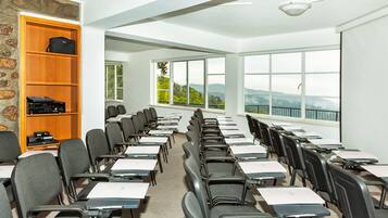 A classroom with rows of desks and chairs, a wooden bookshelf, and large windows offering a view of the outdoors.