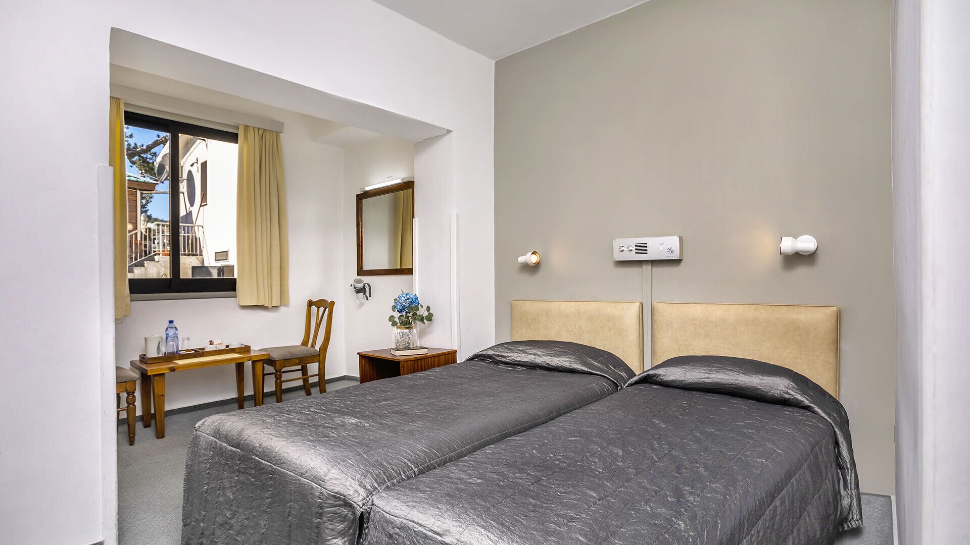 A hotel room with two beds, a desk, a chair, a window with curtains, a mirror, and a wall-mounted light fixture.