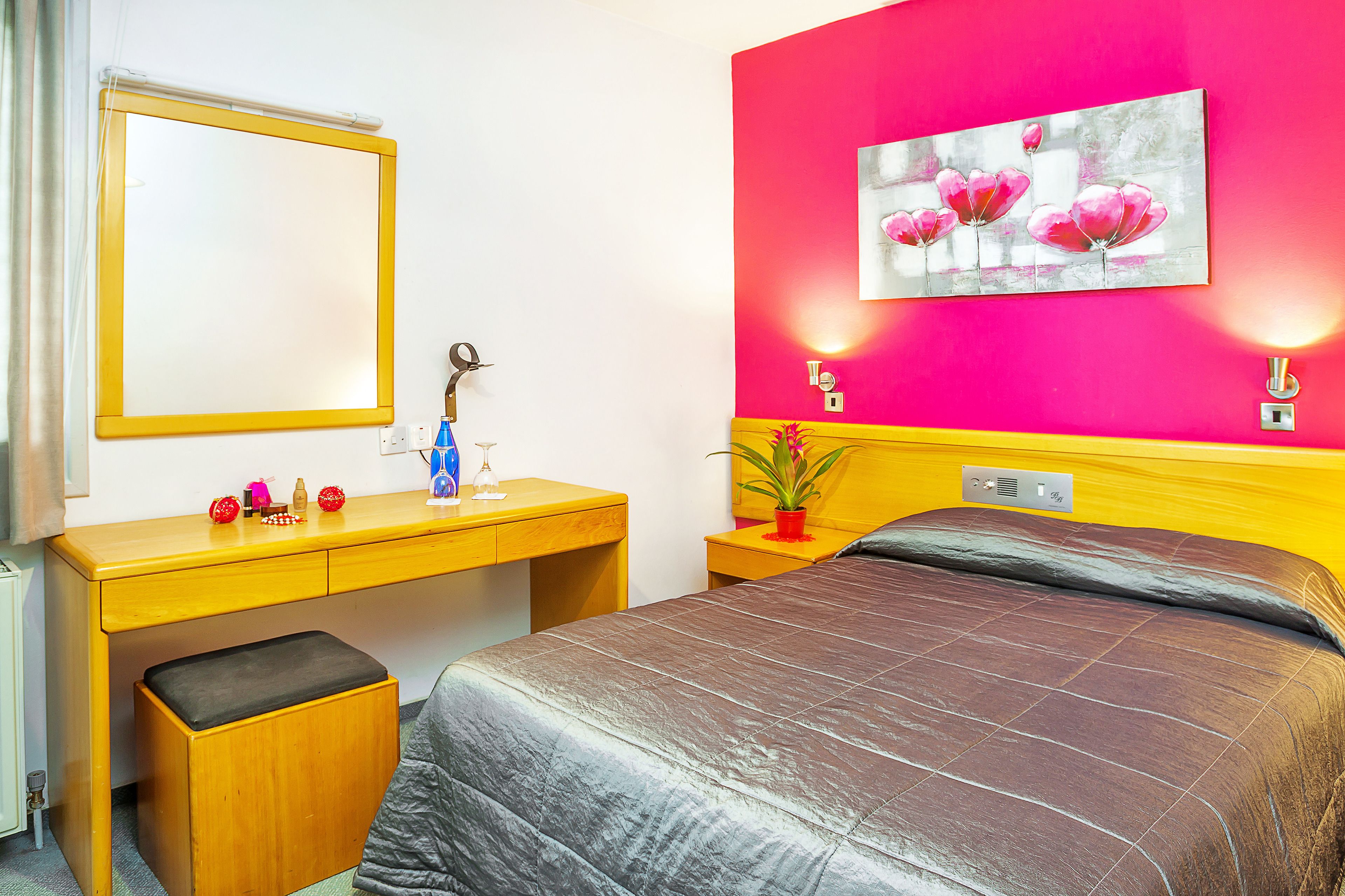 A hotel room with a bed, a desk with a mirror, a small stool, a nightstand with a lamp, and a wall-mounted light fixture.