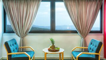 Una habitación con dos sillones azules, una mesa redonda pequeña con un centro de mesa de frutas y vistas a las montañas a través de una ventana con cortinas color beige.