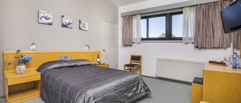 A hotel room with a bed, a wooden nightstand, a chair, a desk, a television, and a window with curtains.