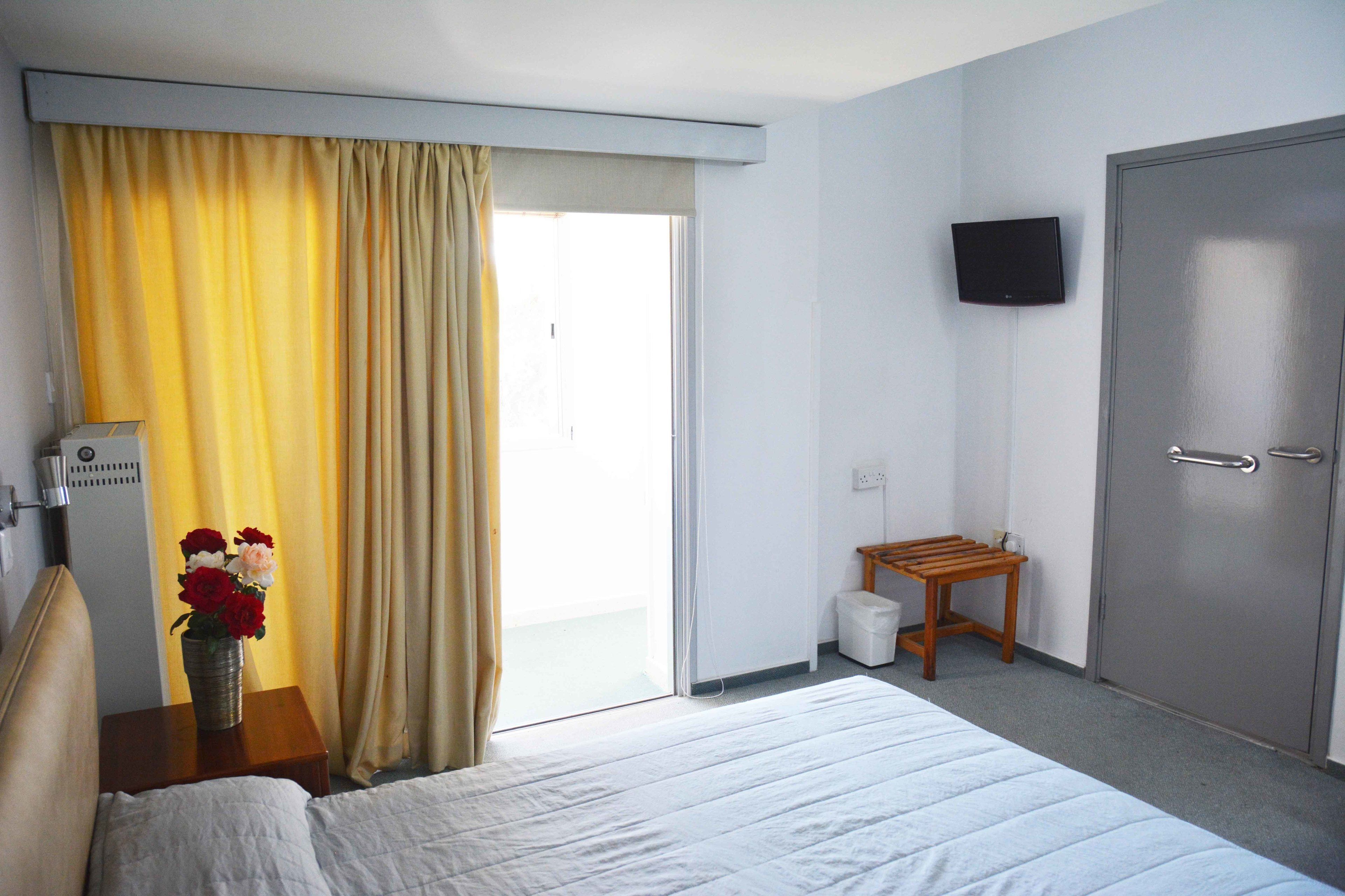 Habitación de hotel con cama, una mesita con un jarrón de flores, televisor montado en la pared y una puerta.