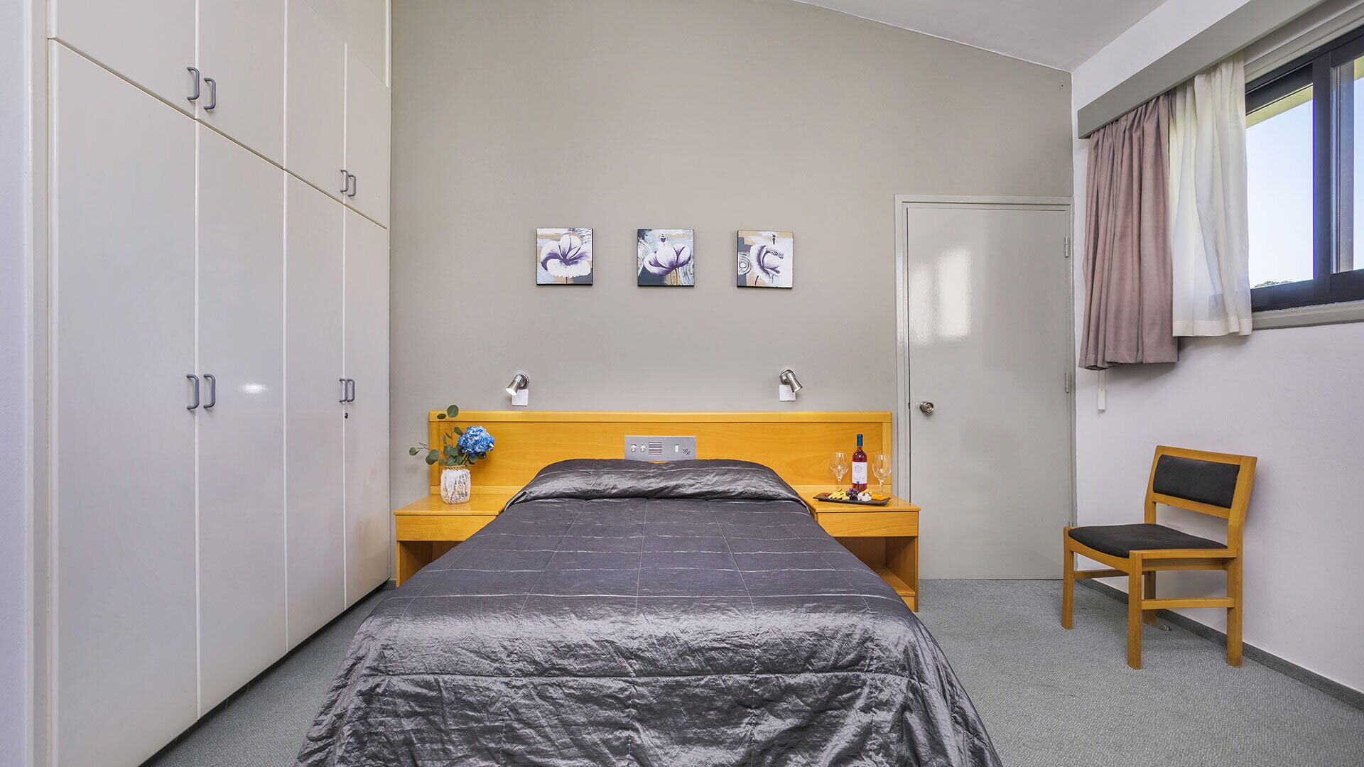 A hotel room with a bed, a wooden nightstand, a chair, a wardrobe, and three paintings on the wall.