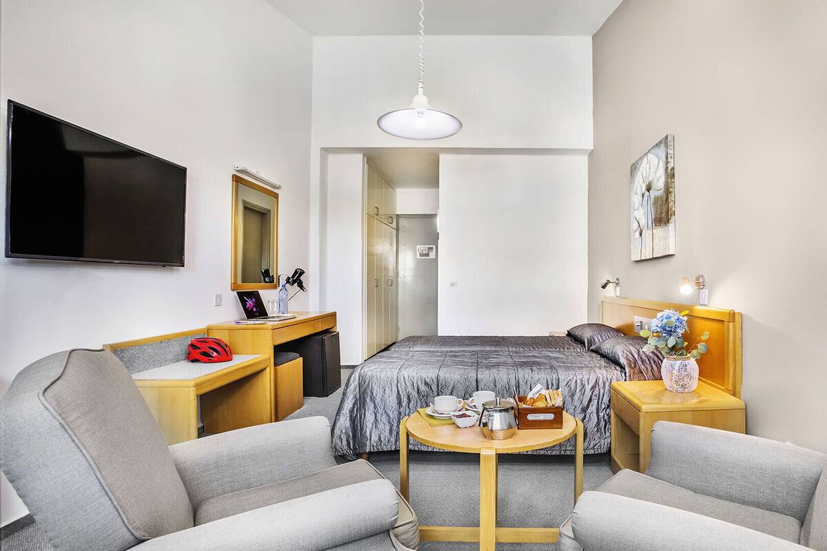Moderni hotellihuone, jossa on sänky, työpöytä, televisio, tuoli, pieni pöytä maljakolla ja taulu seinällä.