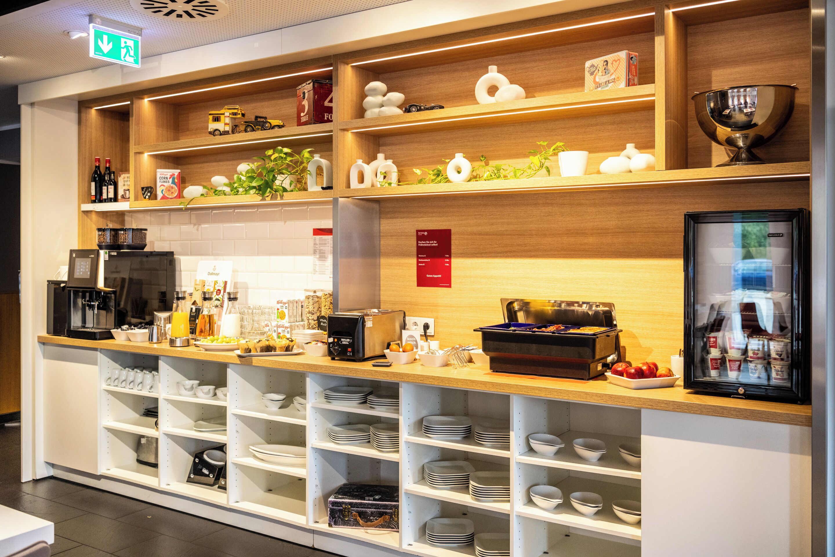 Un área de bufé bien organizada, con diferentes opciones de comida, una estación de café y vajilla dispuesta ordenadamente en estantes.