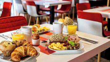 Een ontbijtsetting met croissants, eieren, spek en sinaasappelsap op een tafel in een eetruimte met rode stoelen en tafels.